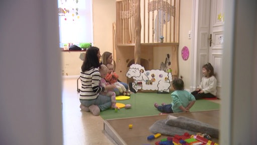 In einer KiTa sind zwei Erzieherinnen und vier Kleinkinder zwischen Spielsachen zu sehen.