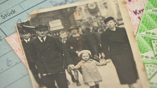 Ein altes schwarz-weiß Foto von einem Kind das in der Mitte an den Händen seiner Eltern geht. 