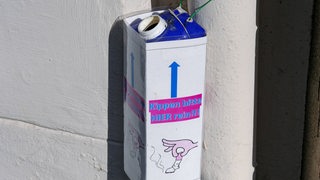Ein Tetrapack hängt an einer Laterne, darauf steht: "Kippen bitter hier rein!" 