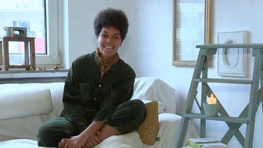 Kira Kayembe sitzt in einer Wohnung gemütlich auf einem Sofa-