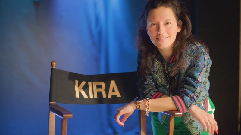 Dunkelhaarige Frau lehnt sich auf Regiestuhl mit Aufschrift "Kira"