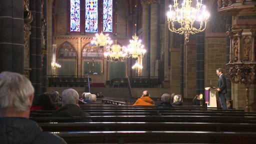 Im Bremer Dom sitzen ein paar Menschen in den Reihen beim Mittagsgebet. Von der Decke hängen Kronläuchter und rechts steht ein Pastor am Rednerpult.