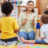 Eine Erzieherin klatscht in die Hände und lacht, Kinder gucken ihr zu