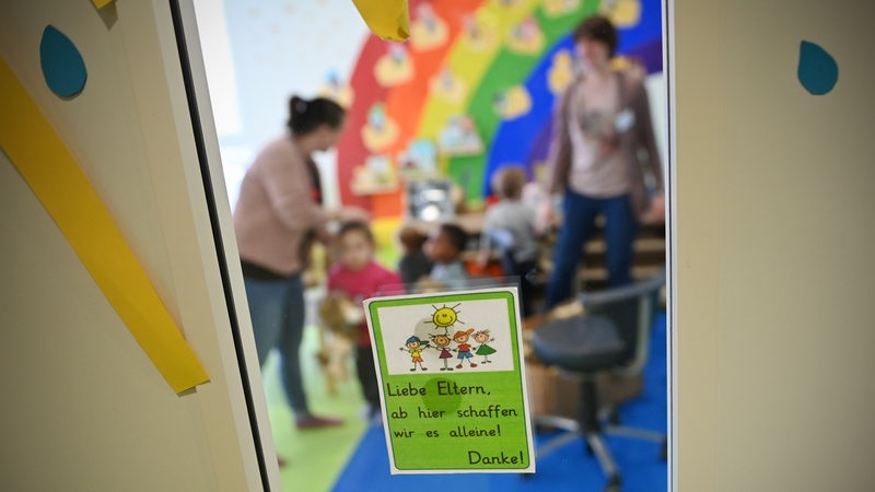 Erzieherinnen mit Kindern in einer Kinderkrippe, vorne an der Tür ein Hinweis an der Tür an die Eltern, draußen zu bleiben (Symbolbild)