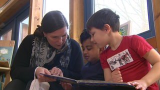 Eine Erzieherin der Kindertagestätte liest zwei Kindern etwas aus einem Buch vor.