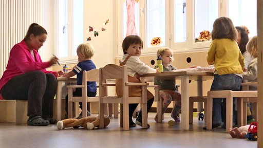 Mehrere Kinder und eine Erzieherin sitzen in einer Kita an kleinen Tischen.