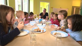 Die Kinder der KiTa sitzen gemeinsam mit ihrer Erzieherin am Mittagstisch.