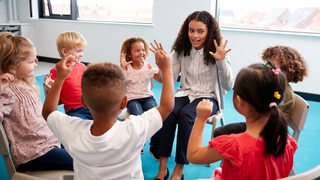 Kinder und eine Frau sitzen in einem Stuhlkreis zusammen und halten die Hände hoch