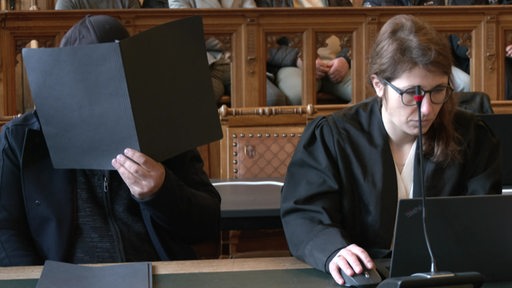 In einem Gerichtssaal sitzt rechts eine Anwältin und links neben ihr ein Angeklagter, welcher sein Gesicht hinter einer Mappe versteckt.
