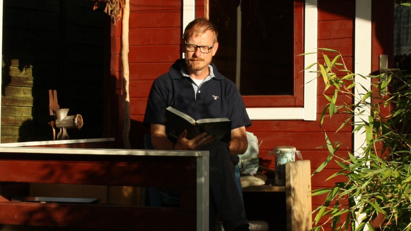Klaus Ferkinghoff sitzt vor einem Blockhaus und liest ein Buch