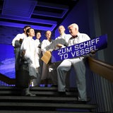 Menschen in weißer Kleidung stehen auf einer Treppe und halten ein Schild mit der Aufschrift "Zum Schiff - to vessel".