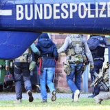 Im Vordergrund ist das Ende eines Hubschraubers zu sehen auf dem Bundespolizei steht. Im Hintergrund wird eine Frau von zwei Polizisten abgeführt. Die drei Personen sind nur von hinten zu sehen und die Frau hat eine Kapuze über ihren Kopf gezogen.