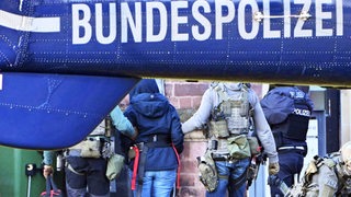 Im Vordergrund ist das Ende eines Hubschraubers zu sehen auf dem Bundespolizei steht. Im Hintergrund wird eine Frau von zwei Polizisten abgeführt. Die drei Personen sind nur von hinten zu sehen und die Frau hat eine Kapuze über ihren Kopf gezogen.