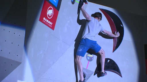 Ein Mann bouldert an einer Kletterwand. Scheinwerfer sind auf ihn gerichtet.