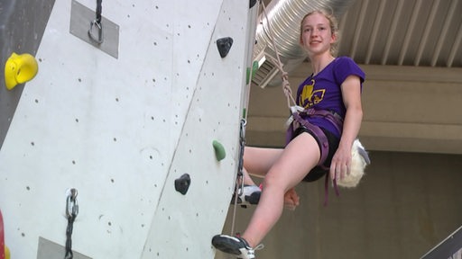 Ein blondes Mädchen hängt abgesichert an einem Seil an einer Kletterwand in einer Sporthalle.