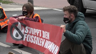 Klima-Aktivistinnen blockieren eine Straße und halten einen Banner, auf dem " STOPPT DEN FOSSILEN WAHNSINN" steht.