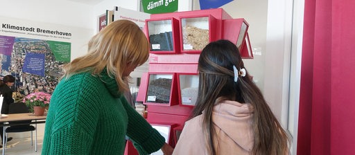 Zwei Personen mit langen Haaren stehen vor einem Aufsteller mit verschiedenen Materialien.