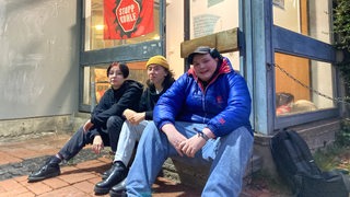 Drei junge Menschen sitzen vor einer Tür mit einem Protest-Banner.