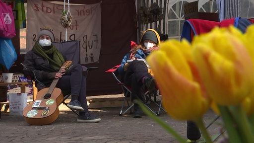 Das Klimacamp in Bremen. Zwei Menschen in Campingstühlen, eine Person hält eine Gitarre. Im Vordergrund ein Strauß Tulpen.