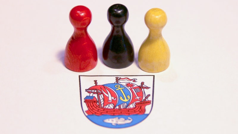 Drei hölzerne Spielfiguren in den Farben Rot, Schwarz und Geld vor dem Wappen von Bremerhaven.