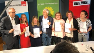 Sieben Personen stehen an einem Tisch und mit dem neuen Koalitionsvertrag zwischen SPD, Grünen und Linken.
