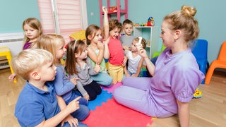 Eine junge Erzieherin sitzt mit einer Kindergruppe auf dem Boden