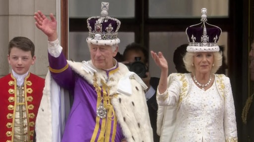 Es sind König Charles und Königin Camilla winkend zu sehen.
