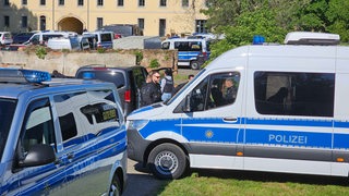 Viele Polizeiwagen stehen vor einem alten Gebäude in Sachsen, Polizeibeamte bewegen sich zwischen den Fahrzeugen.