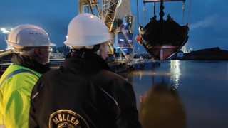Zwei Menschen schauen zu, wie die Hansekogge von einem Kran aus dem Wasser gehoben wird.