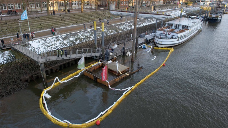 Das Bild zeigt einen Anleger, an dem der Mast und die Dachaufbauten eines Bootes aus dem Wasser schauen.