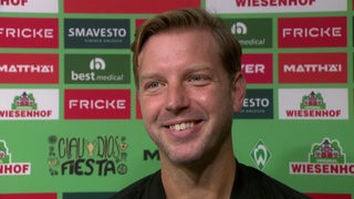 Ex-Werder-Trainer Florian Kohfeldt strahlend vor einer Werbetafel beim Interview.