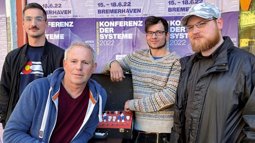 Vier Männer vor drei Plakaten mit Ankündigung zu einer "Konferenz der Systeme" in Bremerhaven