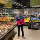 Ein Mann mit einem rosa Einkaufskorb steht zwischen Gemüse-Regalen.