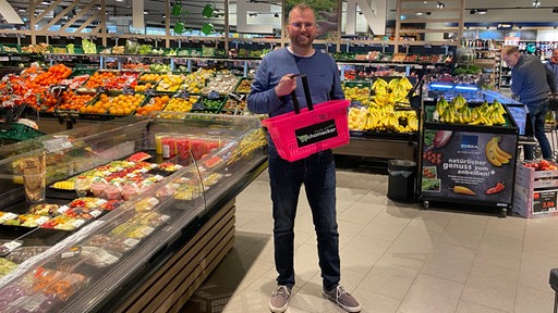 Ein Mann mit einem rosa Einkaufskorb steht zwischen Gemüse-Regalen.