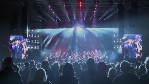 Viele Menschen stehen bei einem Konzert vor einer Bühne mit vielen Künstlerinnen und Künstlern.