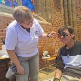 In einer Kirche steht eine Person im weißen T-Shirt vorgebeugt zu einer sitzenden Person, die eine Sehtest-Brille trägt
