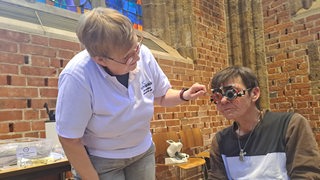 In einer Kirche steht eine Person im weißen T-Shirt vorgebeugt zu einer sitzenden Person, die eine Sehtest-Brille trägt