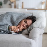 Eine kranke Frau liegt auf der Couch, eingewickelt in eine Decke, und hustet.