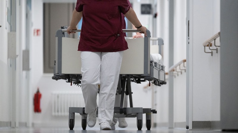 Eine Krankenpflegerin und ein Krankenpfleger schieben ein Krankenbett durch den Gang einer Station eines Krankenhauses.
