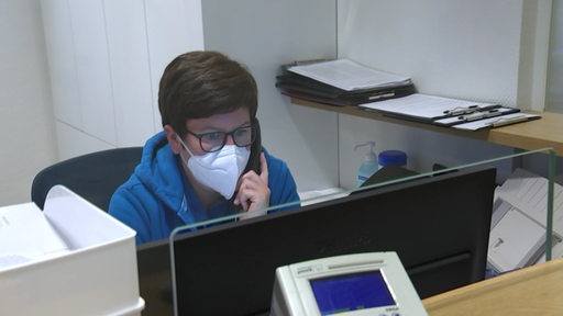 Eine Arzthelferin an ihrem Arbeitsplatz mit Maske und am Telefon. 