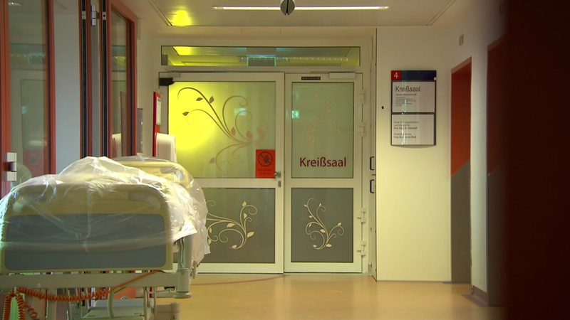 Ein Krankenhaus-Flur, indem ein Krankenbett steht. Am Ende des Flures ist eine Tür, die zum Kreißsaal führt.