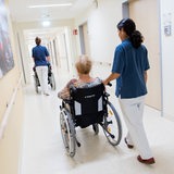 2 Pflegerinnen schieben älteren Patientinnen im Rollstuhl über einen Krankenhausflur.
