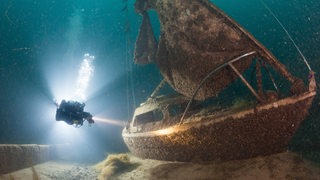 Eine Person taucht vor einem Lichtkegel neben einem Schiffswrack.