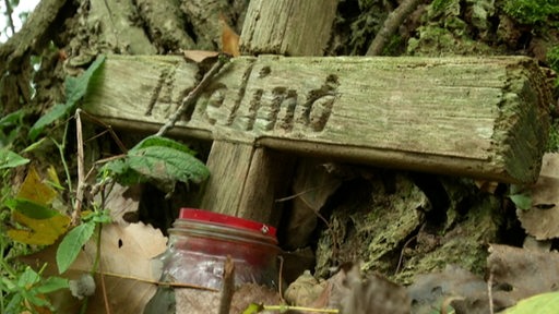 Ein Kreuz aus Holz auf dem Boden mit einer Kerze davor und dem Namen "Adelina" darauf.