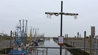 Vor einem Hafenbecken steht ein Kreuz, im Hintergrund liegen Kutter.