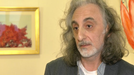 Ein älterer Mann mit Bart und schulterlangen, grau-weißen Haaren schaut nach vorn