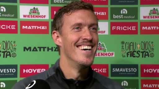 Fußballer Max Kruse steht breit grinsend vor einer Werbetafel beim Interview.