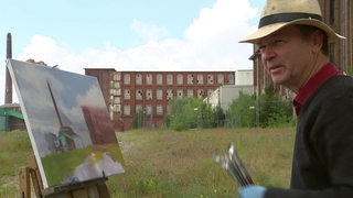 Ein Künstler malt im Freien ein Bild, im HIntergrund sein Motiv. Der Künstler schaut in die Kamera.