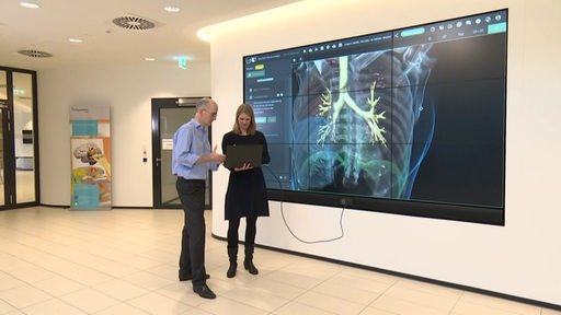 Zwei Personen stehen mit einem Laptop vor einem großen Bildschirm auf dem der Aufbau des menschlichen Körpers abgebildet ist.