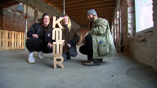 Drei junge Menschen sitzen vor einem Turm aus Buchstaben, die das Wort "Kultur" bilden.